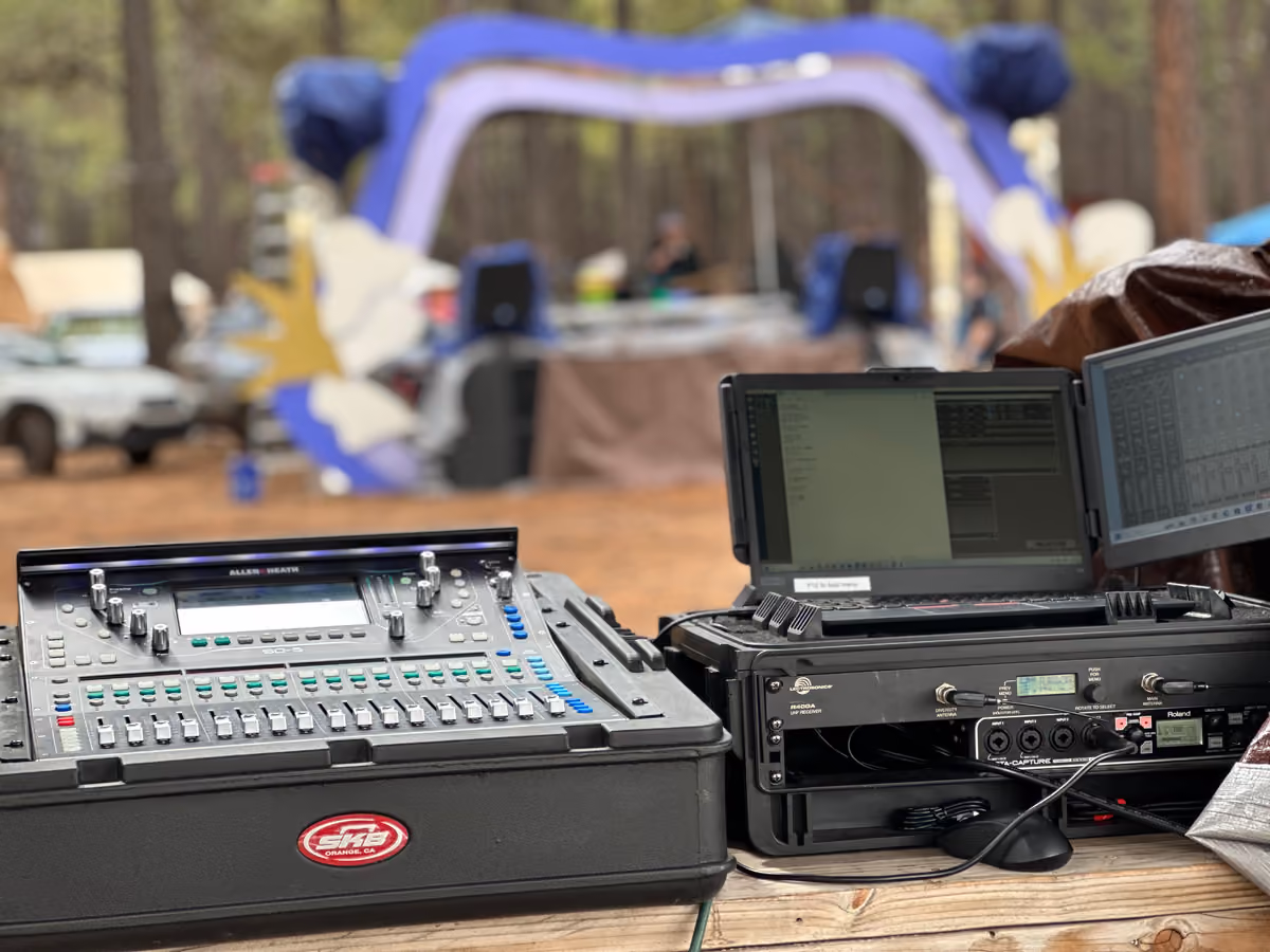 A front-of-house position at a festival in a pine forest: a digital mixing console in a road case, a wireless receiver rack, and a laptop running the system.