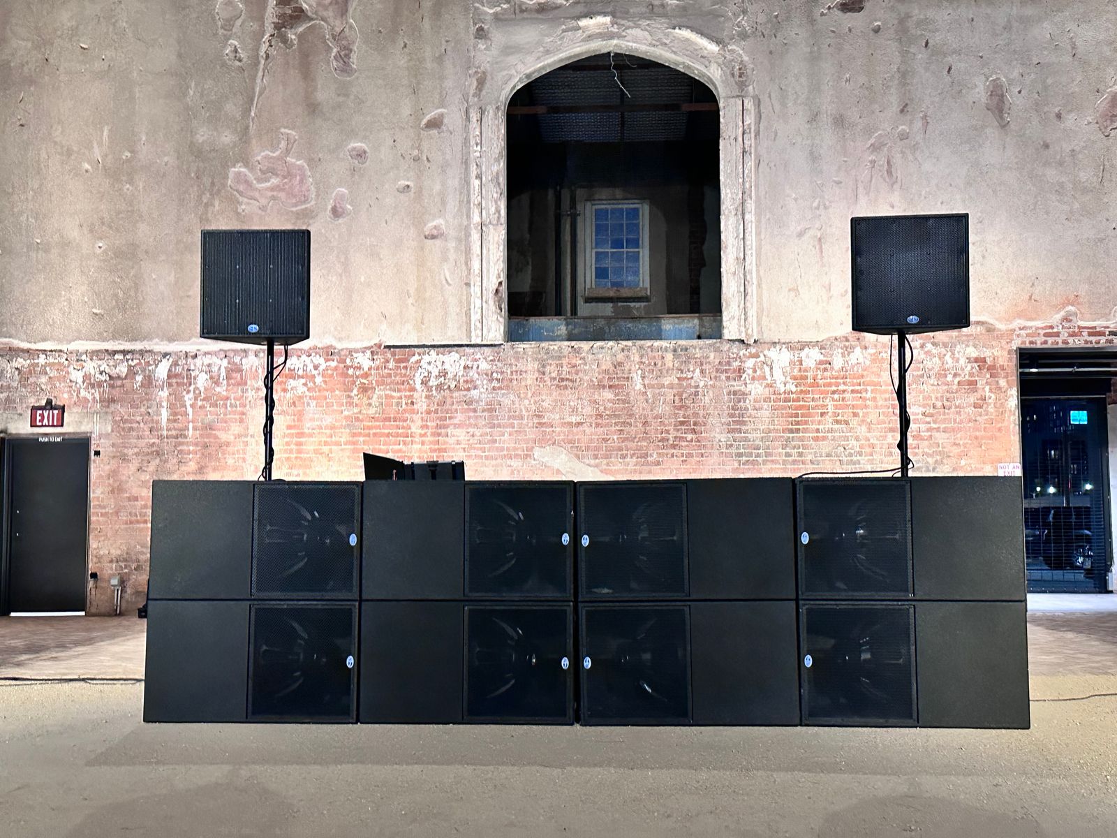 A stacked line array and subwoofers set up against the bare brick wall of a warehouse venue.