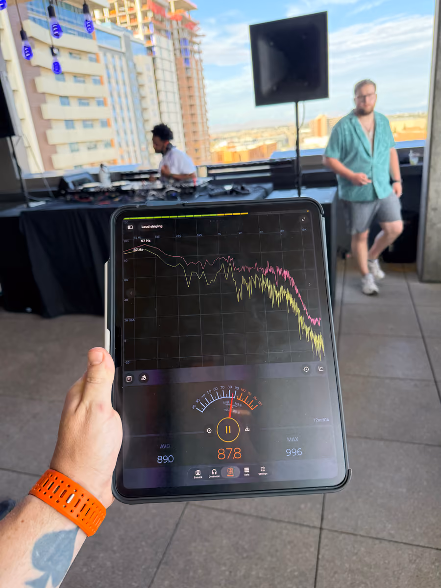 A tablet running a real-time analyzer, showing a frequency response trace and an 87.8 dB reading, held up in front of a rooftop DJ setup.